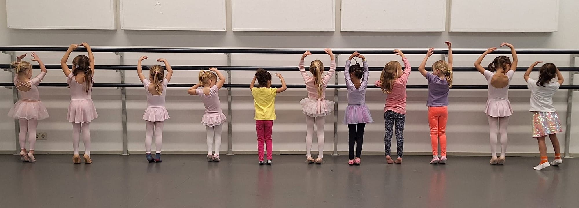 Elf jonge danseresjes aan de barre met de armen in een boog boven het hoofd